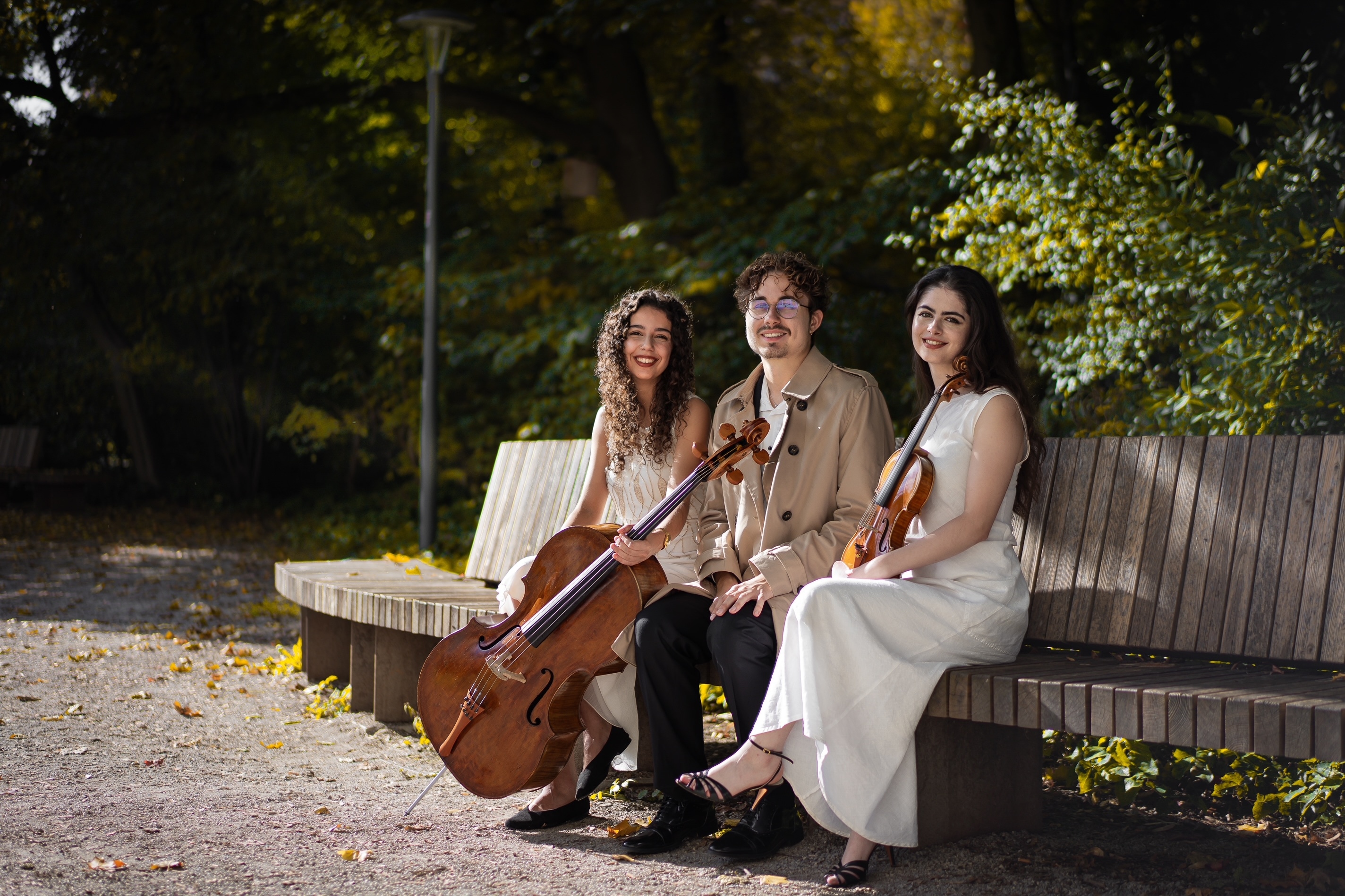 Trio Meran seated outdoors on a curved wooden bench in formal attire, with violin and cello visible.
